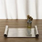 Styled setting with tray on wooden surface in front of white curtain, holding small glass vase with green foliage
