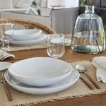 White pasta bowl styled on wooden table with matching plates, clear glass, and decorative glass vase