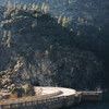 Curved concrete bridge over dark water with steep rocky hillside covered in scattered trees in soft daylight