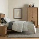 Side view of bed styled in bedroom with neutral linens and matching wood nightstand and wardrobe