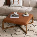 Ottoman placed in living room on patterned rug next to beige tufted sofa with books and cup on top