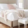 Close-up of bed corner with beige fabric, metallic detailing, and blush throw against wood herringbone flooring