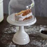 Footed plate holding cake slice under clear glass dome on textured dark wooden surface