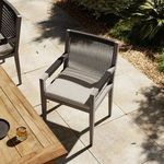 Outdoor patio setting with one chair near wooden dining table, stone tile flooring, shadows from plants nearby