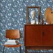 Wallpaper shown in a room setting behind wooden chair and mid-century modern cabinet with decorative items