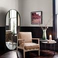 Oval floor mirror placed in room next to beige armchair and side table with decor