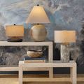 Mythmaker Table Lamp shown on a light-colored textured console table in a room with patterned blue-grey wall