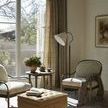 Alternate living room view with painting visible, window light, textured armchairs, and ottoman