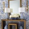 Two lamps flanking square mirror on dark wood console with gold trim, set against blue floral wallpaper