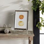 Easel holding a framed art piece with apple prints, displayed on wooden table with decorative objects and plants