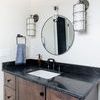 Two sconces mounted on bathroom wall beside round mirror above black granite countertop with dark wood vanity