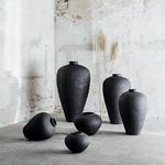 Group of black vases of varying shapes and sizes arranged on concrete floor, with dried grass behind