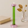 Green and pink glass candle holder lit on a light tabletop with dried leaves and book