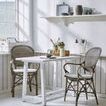 Two Rossini rattan dining armchairs with cushions at small white wooden table with decorative items on shelf above