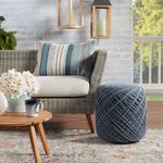 Pillow on wicker chair on patio with round blue knit pouf and wood table with mug and flower vase