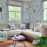 Wallpaper applied in room corner with two windows, beige sofa and brass wall light, showing scale and styling