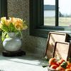 Stoneware vase with yellow tulip flowers on marble counter near basket of oranges and wooden boards