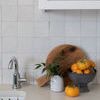 Blue ceramic footed bowl with persimmons displayed on light kitchen countertop near sink and wood cutting board with plant