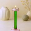 Green and pink glass candle holder with lit candle, displayed on table with blurred decorative objects in background