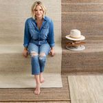 A woman in denim sitting barefoot on the rug against a background of textured woven rugs and hats