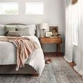 Rug styled in a bright bedroom underfoot beside a bed with light linens and natural wood side table.