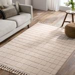 Cream and tan grid rug styled in a living room with beige sofa, wooden floor, round pouf, and side table