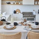 Kitchen setting with black ceramic mugs on white countertop amidst breakfast items and wicker chairs