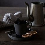 Single black coffee cup on saucer with dried citrus slices, spoon, cloth napkin and vintage metal kettle