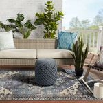 Rug displayed outdoors topped with a blue pouf, surrounded by a wicker sofa and green plants.