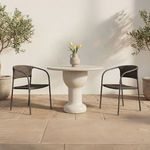Two woven dining chairs next to round cream stone table on textured stone patio flanked by potted plants