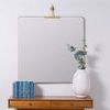 Faith Square Wall Mirror hanging on white wall with wooden table, blue books, and white vase with green foliage