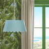 Blue Mirasol Pendant hanging next to cream curtains and a green window frame with ocean view background