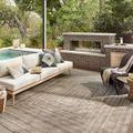 Rug styled outdoors under a pergola with a sofa, pillows, outdoor fireplace, and surrounding greenery