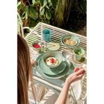 Napkin placed under a green ceramic breakfast set on an outdoor white metal table with plants in background