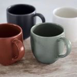 Close-up of artichoke mug among other mugs in black, terracotta, and white on wooden surface