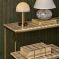 Dezi Box on bottom shelf of brass-framed console with matching travertine tile surface and two tabletop lamps above