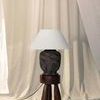 Pecola lamp on wooden tripod stool with visible black cord and cream fabric curtain background