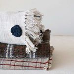 Close-up of textured fringed edges on folded plaid throw with layered neutral throws underneath