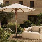 Outdoor setting with Baska umbrella next to curved beige sofa and greenery backdrop