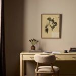 Painting hanging above beige desk with curved chair, books, and vase in softly lit room