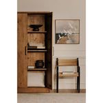 Tade tall cabinet open with decorative items inside, next to woven chair and art on wall