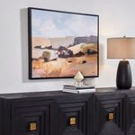 Side angle view of framed desertscape art above black sideboard with lamp and decorative books