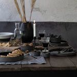 Table set with multiple Lagoa Eco Gres pieces including bowls, plates, and black vase with dried stems under soft lighting
