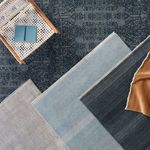 Long narrow blue and ivory rug styled on dark patterned floor with other rugs and wicker chair