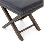 Angled close-up of ottoman corner displaying leather upholstery and brass nailhead row on X frame
