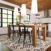 Rug under wood dining table with black chairs in bright kitchen with wood floors and large windows