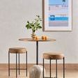 Framed photograph shown hanging on wall above a round wooden table with two leather stools and a vase with green leaves