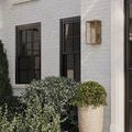 Single antique brass Royster Wall Sconce mounted beside black-framed window and lush green plants