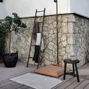 Benji Stool outdoors next to a stone wall, black towel ladder, and wooden outdoor shower