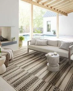 Rug laid out under neutral-toned outdoor sofa on covered patio near swimming pool and fireplace